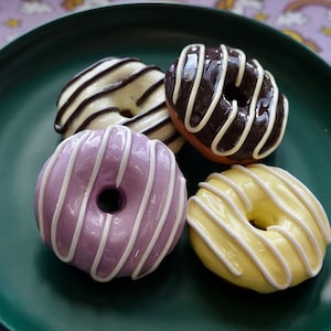 Pode incluir: Quatro donuts coloridos em um prato verde escuro. Os donuts s&atilde;o cobertos com listras brancas sobre coberturas roxas, amarelas, de chocolate e baunilha. O fundo &eacute; um tecido roxo com arco-&iacute;ris e unic&oacute;rnios.