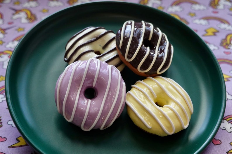 Pode incluir: Quatro donuts coloridos em um prato verde escuro. Os donuts s&atilde;o cobertos com listras brancas sobre coberturas roxas, amarelas, de chocolate e baunilha. O fundo &eacute; um tecido roxo com arco-&iacute;ris e unic&oacute;rnios.