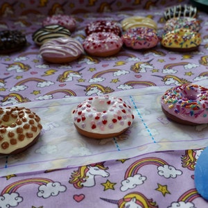 Pode incluir: Uma cole&ccedil;&atilde;o de mini donuts com v&aacute;rias coberturas e decora&ccedil;&otilde;es, incluindo chocolate, granulado e cora&ccedil;&otilde;es. Os donuts est&atilde;o dispostos sobre um tecido estampado com unic&oacute;rnios e arco-&iacute;ris.