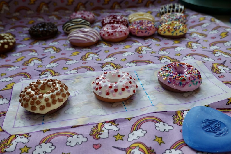Pode incluir: Uma cole&ccedil;&atilde;o de mini donuts com v&aacute;rias coberturas e decora&ccedil;&otilde;es, incluindo chocolate, granulado e cora&ccedil;&otilde;es. Os donuts est&atilde;o dispostos sobre um tecido estampado com unic&oacute;rnios e arco-&iacute;ris.