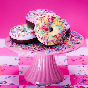 Peut inclure: Trois beignets glacés avec des bases au chocolat et des vermicelles colorés reposant sur un présentoir à gâteaux rose sur pied. Les beignets présentent un glaçage blanc, jaune clair et rose clair, parsemés de vermicelles multicolores.
