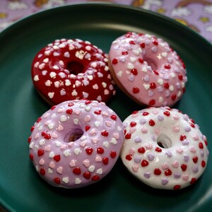 Pode incluir: Quatro donuts decorados com cora&ccedil;&otilde;es em um prato verde escuro. Os donuts t&ecirc;m cobertura rosa, vermelha, roxa e branca, cada um coberto com granulado em forma de cora&ccedil;&atilde;o. O fundo &eacute; um tecido roxo com unic&oacute;rnios e arco-&iacute;ris.
