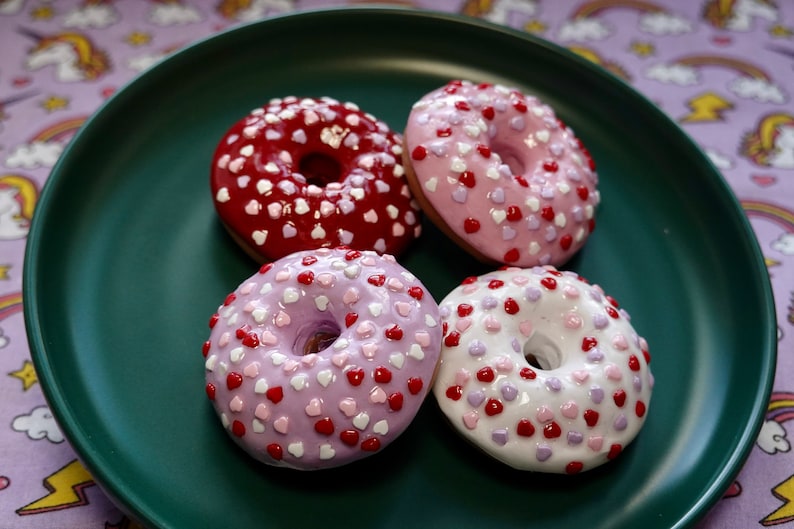 Pode incluir: Quatro donuts decorados com cora&ccedil;&otilde;es em um prato verde escuro. Os donuts t&ecirc;m cobertura rosa, vermelha, roxa e branca, cada um coberto com granulado em forma de cora&ccedil;&atilde;o. O fundo &eacute; um tecido roxo com unic&oacute;rnios e arco-&iacute;ris.