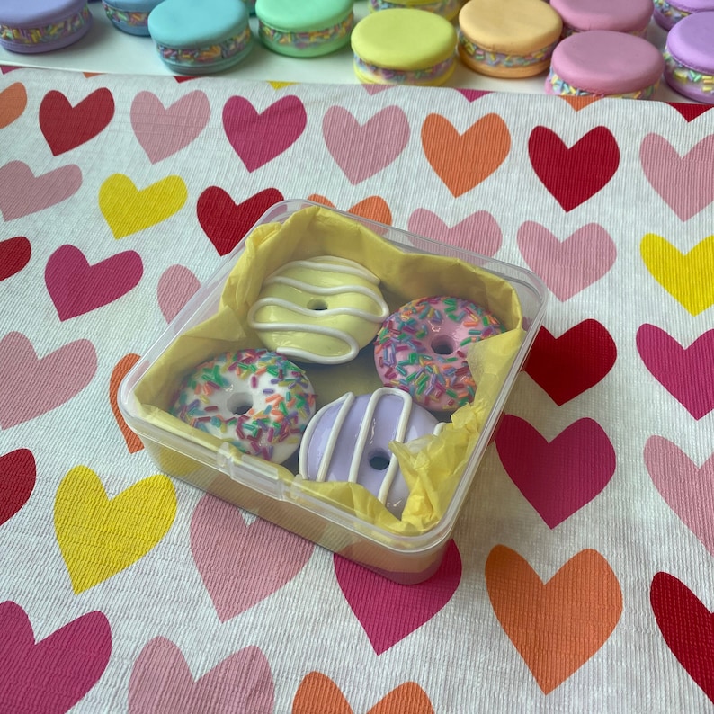 Pode incluir: Um recipiente de pl&aacute;stico transparente cont&eacute;m quatro mini donuts com granulado e cobertura coloridos. Os donuts s&atilde;o amarelos, rosa, brancos e roxos e repousam sobre papel de seda amarelo. O fundo apresenta um tecido com padr&atilde;o de cora&ccedil;&otilde;es e v&aacute;rios macarons.