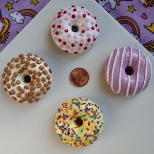Pode incluir: Quatro mini donuts com v&aacute;rias coberturas, incluindo granulado, gotas de chocolate e glac&ecirc;. Uma moeda de um centavo est&aacute; no centro. Os donuts est&atilde;o dispostos em uma superf&iacute;cie branca com um fundo roxo com tema de unic&oacute;rnio.