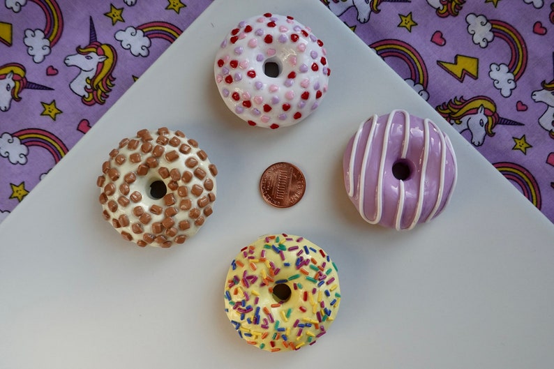 Pode incluir: Quatro mini donuts com v&aacute;rias coberturas, incluindo granulado, gotas de chocolate e glac&ecirc;. Uma moeda de um centavo est&aacute; no centro. Os donuts est&atilde;o dispostos em uma superf&iacute;cie branca com um fundo roxo com tema de unic&oacute;rnio.