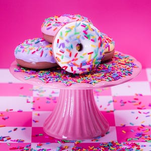 Puede incluir: Una pila de tres donas glaseadas cubiertas con chispas de colores sobre un soporte para pasteles rosa con pedestal. Las donas presentan glaseado blanco, morado claro y rosa, cubiertas de chispas multicolores, con chispas sueltas en el soporte.