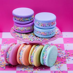 Puede incluir: Una colección de galletas sándwich redondas, lisas y de colores pastel con un relleno de crema blanca y virutas de colores. Dos galletas están apiladas en un soporte rosa, mientras que otras cuatro están dispuestas verticalmente debajo.