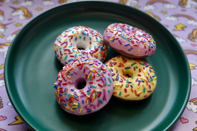 Pode incluir: Quatro donuts coloridos com granulado num prato verde escuro. Os donuts t&ecirc;m cobertura branca, rosa, roxa e amarela. O granulado &eacute; multicolorido. O prato est&aacute; sobre uma toalha de mesa roxa com um padr&atilde;o de unic&oacute;rnios.