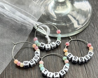 May include: Four silver hoop wine charms with white letter beads spelling "WILD", "WOOP", "TIPSY", and "SOBER". The charms have pastel-colored beads and are near a wine glass.
