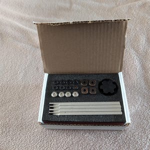 May include: An open cardboard box containing various small, black, brown, and silver metal components. The components are arranged on a white foam insert. The box is on a beige surface.