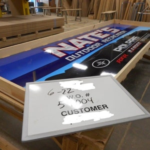 May include: A wooden frame with a blue and white sign that says "Nate's Outdoor Recreation" with a Polaris RZR Ranger logo. A white board with the text "6-22 W.O. # 51004 CUSTOMER" is leaning against the frame.