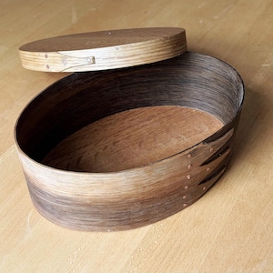 May include: An oval Shaker box made of wood, with a lid slightly ajar. The box features a gradient effect on the sides, transitioning from light to dark brown. A traditional craft item, the box is designed for storage.