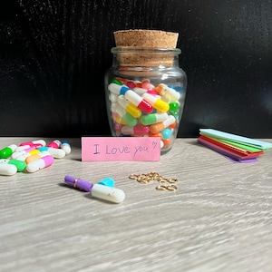 Op de afbeelding: Een glazen pot gevuld met kleurrijke capsules, elk met een smiley. Een roze plakbriefje met de tekst "I Love you" ligt naast de pot. Er liggen ook verschillende kleine capsules en gouden springringen op de tafel.
