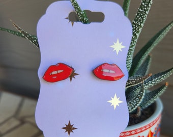 Könnte beinhalten: Ein Paar rote Lippen-Ohrstecker auf einer lila Karte mit goldenen Sternen. Die Ohrringe befinden sich auf einem weißen Hintergrund.