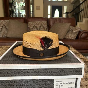 May include: A butterscotch-coloured Panama hat with a black band and a decorative feather. The hat has a 6 cm brim and is size 7. The hat is presented on a box featuring the Stetson Legendary Hats logo.