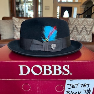 May include: A black fedora hat with a decorative feather and a ribbon band. The hat has a 1 7/8 inch brim and is size 7 7/8. The hat is on a red box with the brand name "DOBBS" in white.