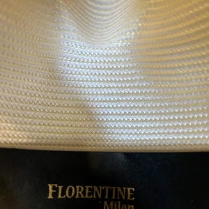 Puede incluir: Primer plano del interior de un sombrero fedora color crema. La textura tejida del sombrero es visible, con las palabras "FLORENTINE Milan" en letras doradas sobre una banda de cuero negro. El interior del sombrero es de color crema claro.