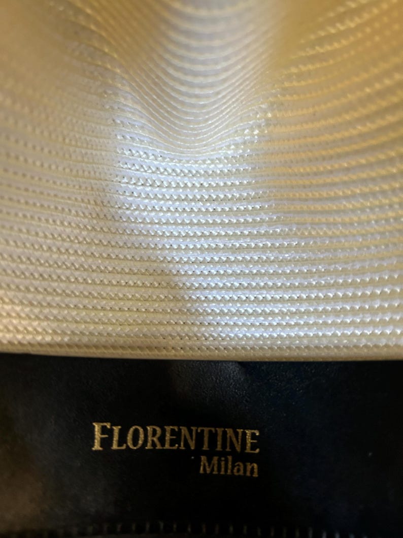 Puede incluir: Primer plano del interior de un sombrero fedora color crema. La textura tejida del sombrero es visible, con las palabras "FLORENTINE Milan" en letras doradas sobre una banda de cuero negro. El interior del sombrero es de color crema claro.