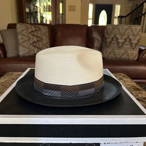 Puede incluir: Un sombrero fedora Stetson marfil/negro con un ala de 5,7 cm. El sombrero presenta una corona color crema, un ala negra y una banda estampada con detalles marrones y grises. El sombrero se exhibe en una caja negra.