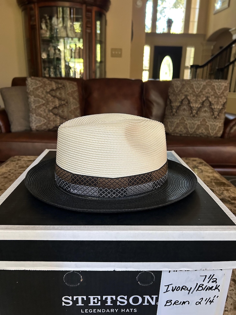 Puede incluir: Un sombrero fedora Stetson marfil/negro con un ala de 5,7 cm. El sombrero presenta una corona color crema, un ala negra y una banda estampada con detalles marrones y grises. El sombrero se exhibe en una caja negra.