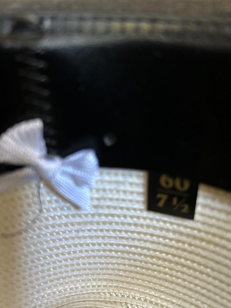 Puede incluir: Primer plano del interior de un sombrero de paja. El sombrero presenta una cinta blanca y una etiqueta que indica la talla 60 y 7 1/2. El interior del sombrero es blanco con una textura tejida y el ala es negra.