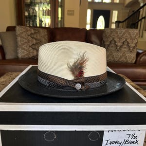 Puede incluir: Un sombrero fedora Stetson color crema con ala negra y pluma decorativa. El sombrero est&aacute; sobre una caja en blanco y negro. El sombrero tiene una banda marr&oacute;n y gris con un emblema plateado.