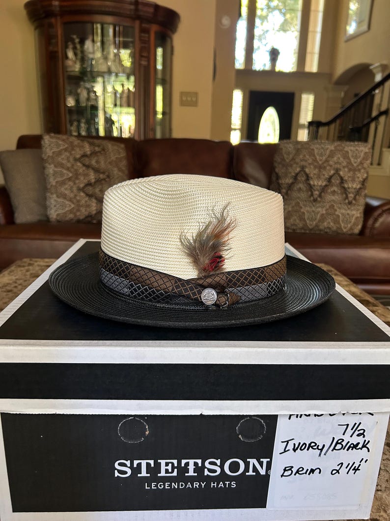 Puede incluir: Un sombrero fedora Stetson color crema con ala negra y pluma decorativa. El sombrero est&aacute; sobre una caja en blanco y negro. El sombrero tiene una banda marr&oacute;n y gris con un emblema plateado.