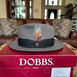 May include: A grey fedora hat with a black band and decorative feather. The hat is displayed on a red box with the brand name "DOBBS" in white. The box also has product details, including "GOLD COACH 85 Steel".
