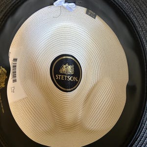 Puede incluir: Vista interior de un sombrero fedora Stetson Florentine Milan negro. El sombrero presenta una corona tejida de color crema y una banda de sudoraci&oacute;n de cuero negro. El logotipo de Stetson es visible en una etiqueta negra y dorada.