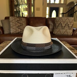 Puede incluir: Un sombrero fedora Stetson marfil y negro con un ala de 5,7 cm, sobre una caja de sombreros negra. El sombrero presenta una corona color crema, una banda marr&oacute;n y un ala negra. La caja del sombrero tiene el logotipo de Stetson.