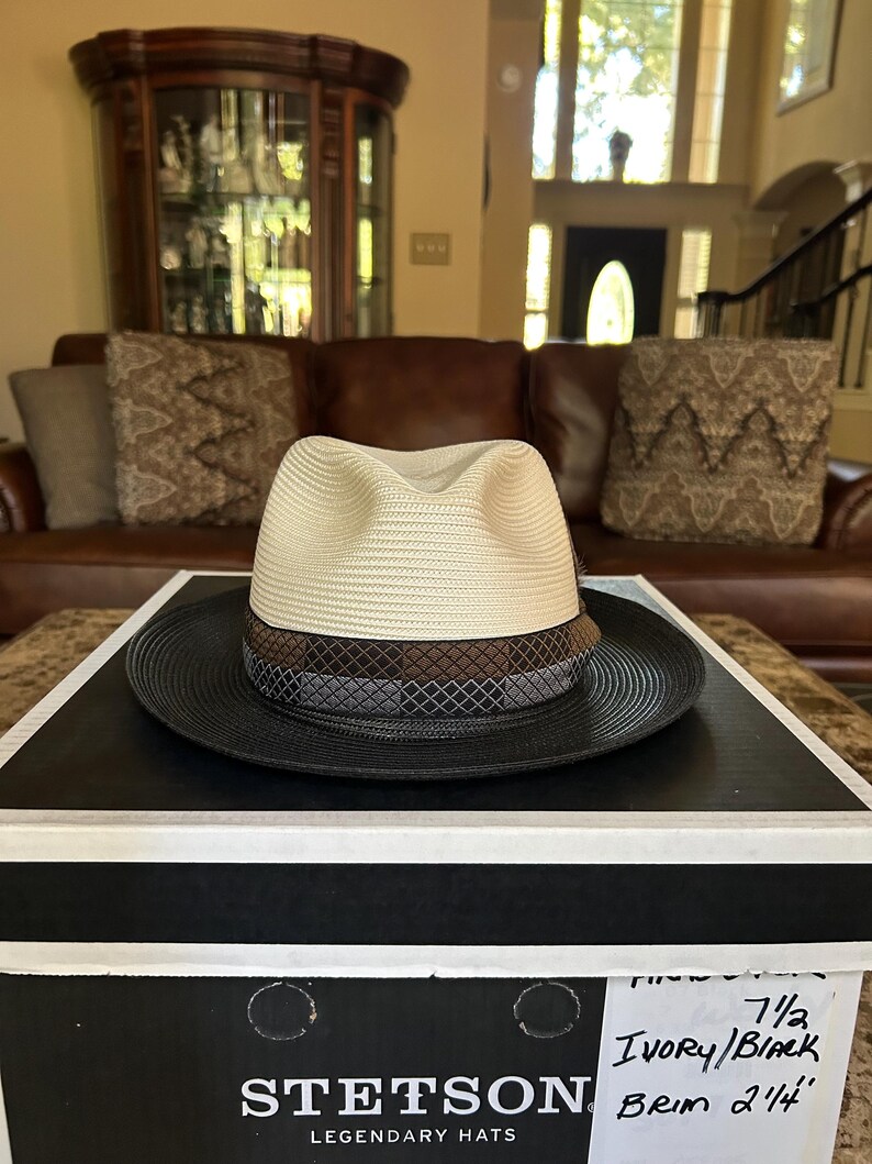 Puede incluir: Un sombrero fedora Stetson marfil y negro con un ala de 5,7 cm, sobre una caja de sombreros negra. El sombrero presenta una corona color crema, una banda marr&oacute;n y un ala negra. La caja del sombrero tiene el logotipo de Stetson.
