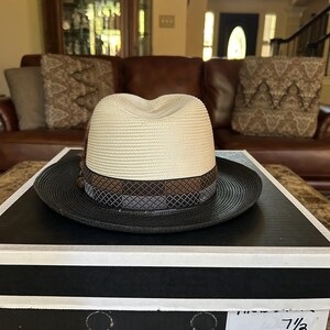 Puede incluir: Sombrero fedora Stetson marfil y negro con ala de 5,7 cm, sobre una caja en blanco y negro. El sombrero tiene una corona crema, una banda marr&oacute;n y gris y un ala negra. La caja muestra el logotipo de Stetson.