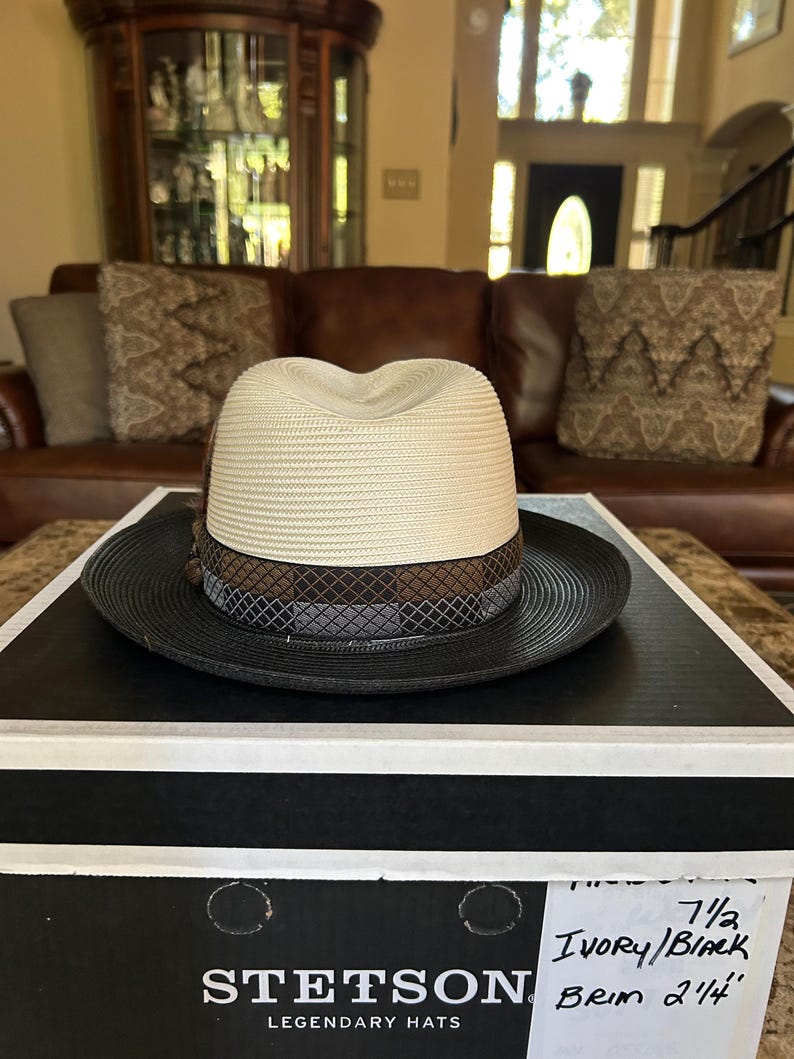 Puede incluir: Sombrero fedora Stetson marfil y negro con ala de 5,7 cm, sobre una caja en blanco y negro. El sombrero tiene una corona crema, una banda marr&oacute;n y gris y un ala negra. La caja muestra el logotipo de Stetson.