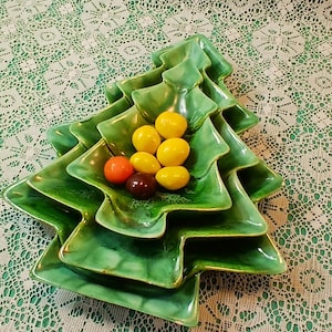 May include: A green ceramic Christmas tree-shaped serving dish, stacked in tiers, filled with colorful candies. The dish is on a white lace tablecloth. A festive holiday serving piece.