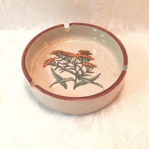 May include: A round ceramic ashtray with a speckled cream-colored base and a dark brown rim. The center features a detailed illustration of a cluster of orange-flowered plants with green stems and leaves. The ashtray has two notches.