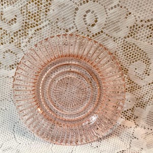 May include: A round, pale pink glass saucer featuring a textured, pressed pattern. The design includes a central circular motif with a grid-like texture, surrounded by a smooth ring and an outer border of vertical, radiating fluted ridges.