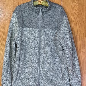 May include: A full-zip fleece jacket featuring a two-tone gray color scheme. The upper chest and collar are a solid dark gray, while the lower body and sleeves are a lighter gray with a speckled, textured pattern. It includes a vertical zippered chest pocket and two lower zippered side pockets.