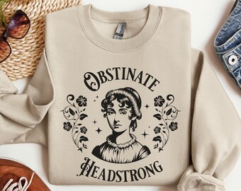 May include: A beige sweatshirt with a black graphic print of a woman's face with the text "Obstinate Headstrong" in a circle with floral accents.