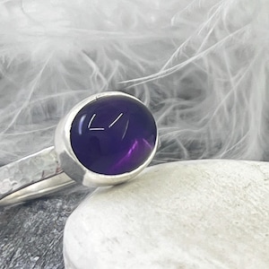 Puede incluir: Un anillo de plata con una gema grande, redonda y de color púrpura intenso. La banda tiene una textura martillada. La gema está engastada en un bisel liso. El anillo se muestra sobre una superficie blanca con un fondo suave y borroso.