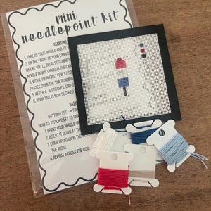 May include: A mini needlepoint kit with instructions and a partially stitched design of a red, white, and blue buoy. The kit includes a canvas, black frame, needle, and spools of red, white, blue, and tan thread. The kit is on a wooden surface.