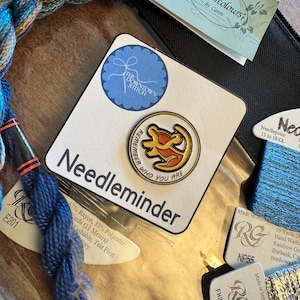 May include: A collection of embroidery supplies, including blue and multicolored thread, a "Needleminder" with text and a circular design, and tags from "Watercolours by Caron" and "The Downtown Stitch."