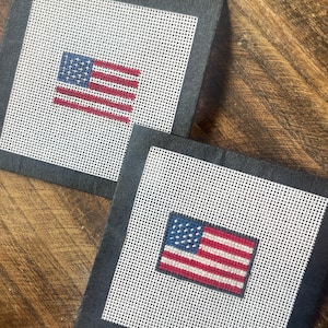 May include: Two square coasters with a black border and a white mesh center. Each coaster features a stitched American flag design with red and white stripes and a blue field of stars. The coasters are displayed on a wooden surface.