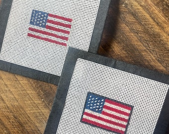 Puede incluir: Dos posavasos cuadrados con borde negro y centro de malla blanca. Cada posavasos presenta un diseño de bandera estadounidense cosida con rayas rojas y blancas y un campo azul de estrellas. Los posavasos se muestran sobre una superficie de madera.