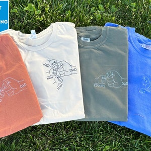 May include: Four t-shirts with a fist bump design embroidered on the front. The shirts are in different colors: orange, beige, green, and blue. The fist bump design includes the names "Ted, Dad", "Mike, Karlie, Brian, Dad", "Rodney, Dad", and "Lily, Jack, Dad".