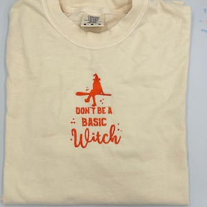 May include: A white t-shirt with an orange embroidered design of a witch on a broomstick. The text "Don't be a basic witch" is also embroidered in orange.