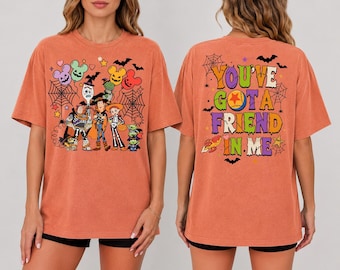 Può includere: Una t-shirt arancione a maniche corte con personaggi di Toy Story in costumi di Halloween sul davanti e il testo "YOU'VE GOT A FRIEND IN ME" con una grafica a palla sul retro. Il design include ragnatele, pipistrelli e palloncini colorati.