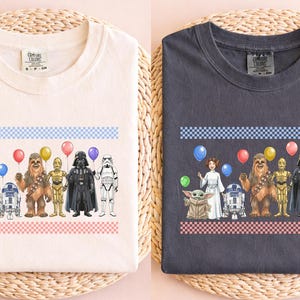 May include: Two t-shirts, one white and one charcoal gray, each featuring a colorful graphic of Star Wars characters holding balloons. The design includes characters like Yoda, Darth Vader, and Chewbacca, with a blue and pink checkered border.
