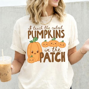 May include: An ivory short-sleeve t-shirt featuring a brown graphic print that reads I teach the cutest PUMPKINS in the PATCH. The design includes five orange cartoon pumpkins with smiling faces and green stems arranged in a cluster on the front.