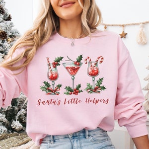May include: A light pink sweatshirt with a Christmas-themed design featuring three cocktails with holly and candy cane accents. The text "Santa's Little Helpers" is printed below the cocktails.
